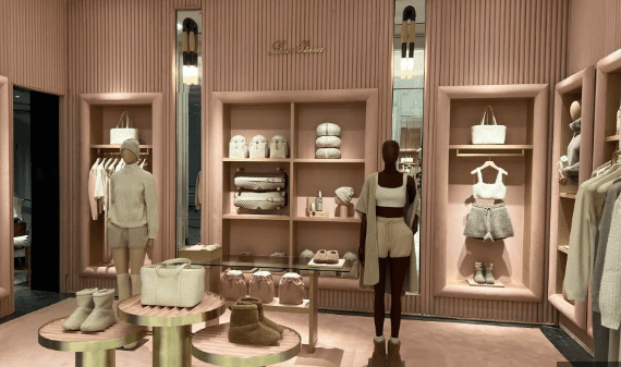 Interior of a Loro Piana luxury boutique installation, possibly at Bergdorf Goodman, featuring a monochromatic palette of blush pink and beige. Mannequins display cashmere loungewear and accessories on plush, gold-accented displays.